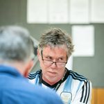 An older white man with shaggy hair on top is sat he is looking over black rimmed glasses to his left. He has light white stubble. His arms are folded but half of his body is obstructed by another man with his back to the camera wearing a blue shirt.