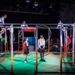 Two cast members in the centre of the stage as the others look down them from the perimeter metal frame of the set.