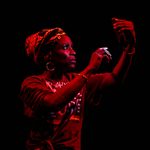 Middle-aged Black woman in red lighting, holding a phone and staring her phone screen, wearing a colorful headdress and gold hoop earrings.