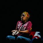 A middle-aged Black woman, adorned with a vibrant headdress and gold hoop earrings, captured in a mid-action shot while sitting on a Union Jack flag-covered chair, her face showing a deeply expressive and emotional expression.