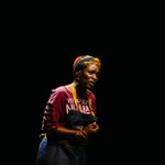 A middle-aged Black woman with a colorful headdress and gold hoops, displaying a pained and anguished expression.