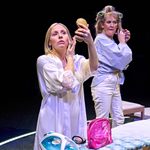 Two women stand getting ready, one wears a white dressing gown