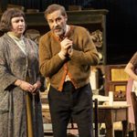 Siobhán McSweeney as Maggie wearing a long grey floral cardigan standing next to Ardal O'Hanlon as Jack in a brown cardigan. Justine Mitchell as Kate watches them in a purple dress.