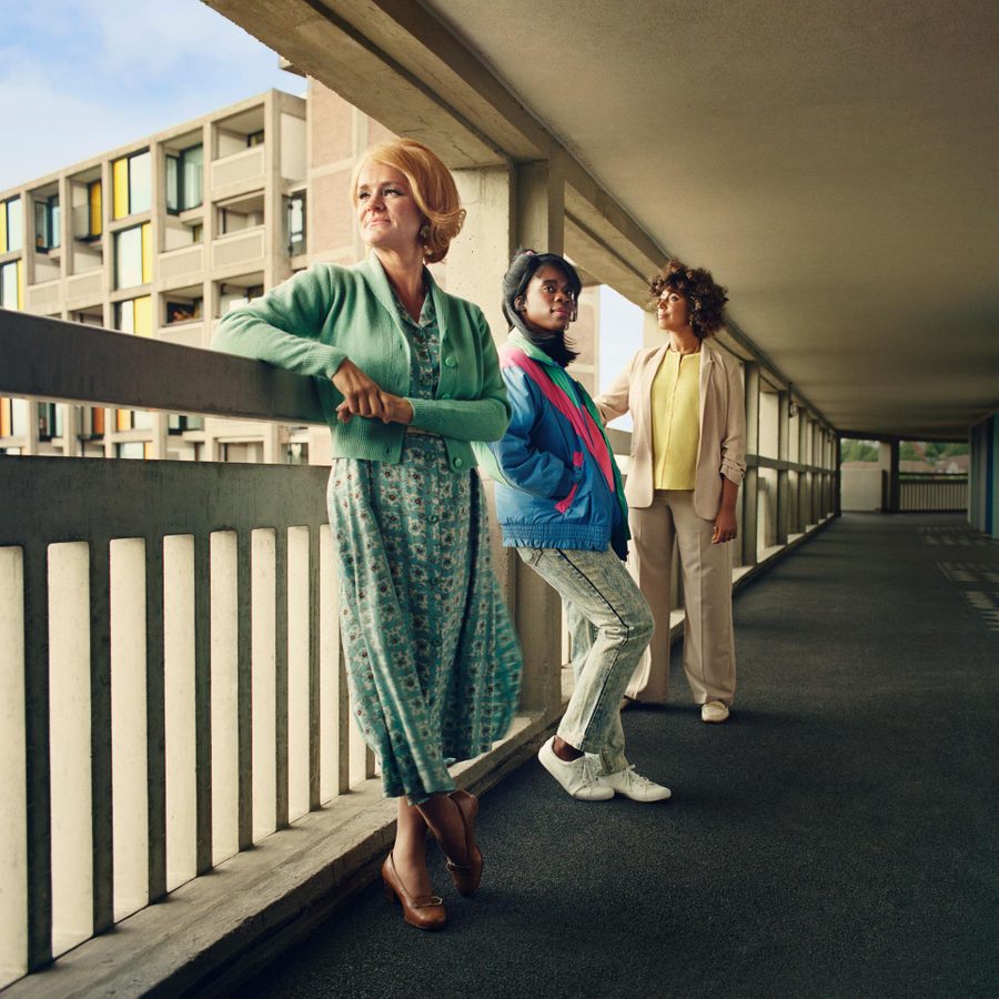 Three women, of mixed age and race, leaning against railings on a walkway in a block of flats.