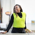 A smiling woman, wearing a bright yellow, sleeveless knitted top over a black jumper, stands, leaning against a kitchen work-top.