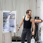 A man wearing a black vest and jeans, stands looking away from a fridge-freezer with eth upper fridge door open. An electric heater on a stand is behind him.