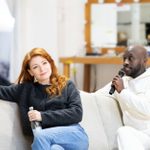 A woman holds a wine glass and bottle, a man speaks into a microphone. They are both sitting on a plain, off-white coloured sofa.