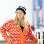 A woman stands, leaning against a kitchen work-top. She wears a knitted had and brightly coloured cardigan.