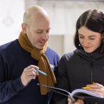 A man and woman stand side by side, looking at open pages in a binder. Both are smiling and holding pens.