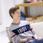 A young man wearing a top with 1891 printed on it, sits on a plain, off-white coloured sofa.