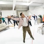 A man stands in a white room, leaning to his right, stretching our his arm and a group of people behind him copy his movement.