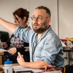 Stephen Brackett, sitting at a desk and gesturing with right arm extended, in rehearsals for A Strange Loop