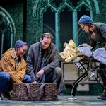 Three people stand on stage staring down at a wicker baby basket. A large dog-like puppet made of paper is peering into the basket too, with the puppeteer behind him dressed in plain clothes.