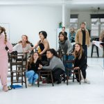 Cast members standing, seated or crouching on and around a number of wooden chairs, acting a scene in a large white room.