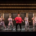 A character in a red bell-hop cap and jacket dances centre-stage, with a line of 6 other characters behind him