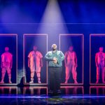 A character in a long-sleeves shirt stands centre-stage, in a spotlight, with a line of 6 other characters behind him, all standing in a doorway framed in neon strip-lights.