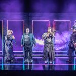 Seven characters in a variety of outfits stand across a stage lit in blues and with smoke swirling upstage.