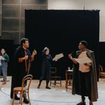 Six people in rehearsals. Three people are wearing old travelling cloaks over their casual clothes, four are holding smoking pipes, and two are holding newspapers. There are various chairs throughout the space.