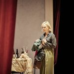 A young woman stands next to table with a rotary phone on is. She is holding a green coat and clutching a black leather handbag. She has a blonde bob and is swearing a green shirt which has pink diamond pattern, she is also wearing a green skirt