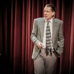 A older man stands against a red theatre curtain. He has long hair scraped back, he wears a grey suit with a patterned tie.