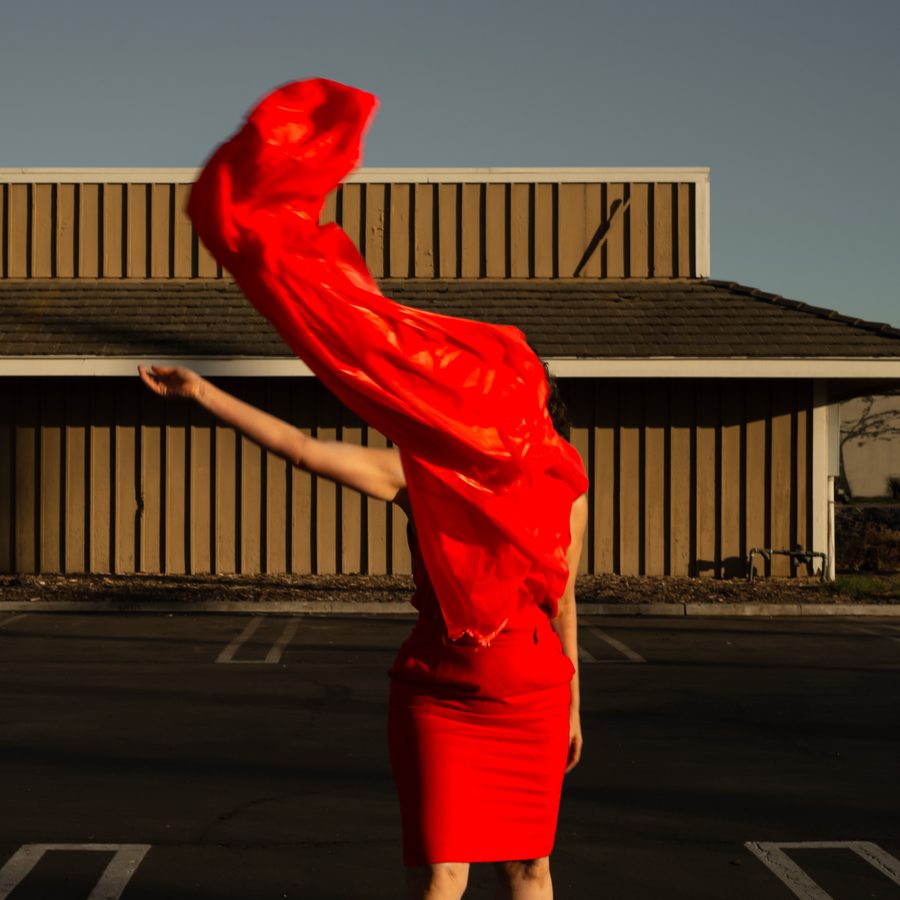 A figure with one arm outstretched, their body and head wreathed in billowing red material, stands in front of a low-rise building with no doors or windows.