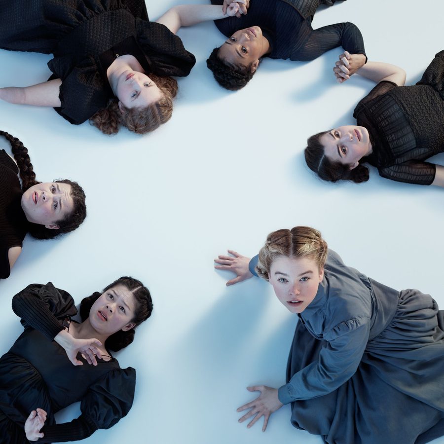 Three young women lying on a white floor, all looking up, two holding hands are on the left of the image, the third pushing herself up on her hands, on the right of the image.