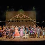 A large group of people onstage singing with their arms outstretched with garlands and bunting hung above them.