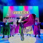 A group of performers onstage, dressed in sequin outfits, singing with a big sign saying 'JUSTICE' hanging above them.