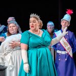 In the foreground, a woman dressed in a tiara and turquoise dress speaking. In the background, a woman holding a baby and a man dressed as a palace guard.