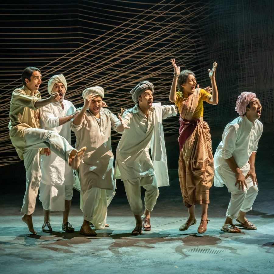 Seven people wearing loose cotton clothes, standing in a line. in celebratory poses.