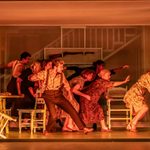 A large group of people lurch towards two women holding each other. They are in a house which is lit by orange light.