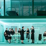 Thirteen women stand and sit in a house. The house's walls and furniture are all the same blue-green colour.