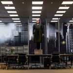 A white man wearing a black 3-piece suit and round glasses stands on a table in a glass office. A white man stands in front of the table to his left, and a black man stands to his right. Both are wearing black 3-piece suits and looking up at the man on the table. There are projections of the New York skyline in the background.