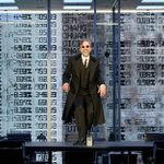 A white man wearing a black 3-piece suit and black sunglasses dances on a table in a glass office. There are projections of numbers and percentages behind him.