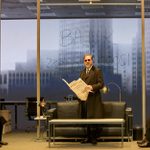 A white man wearing a black 3-piece suit and black sunglasses stands in a glass office holding a newspaper. A black man sits on his left and a white man sits on his right. Behind them the word BANK and some numbers are written on the glass wall and there are projections of the New York skyline in the background.