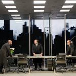 A black man and two white men wearing black 3-piece suits stand around a table in a glass office, staring intently at each other. There are projections of the New York skyline in the background.