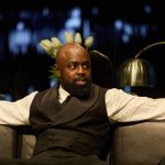 A black man wearing a white shirt and black waistcoat sits on a sofa in an office. Behind him there is a silver desk lamp and a bunch of white tulips.