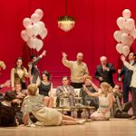 A group of people are having a party in a large room with red panelled walls and cream furniture and floor. Some are standing, some sit on the sofa and chairs and some lie or kneel on the floor. There are two large bunches of white balloons.