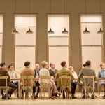 Thirteen people are sitting in hard-backed chairs around a long table, all looking towards a man who stands at one end, pointing to the man at the other end of the table. The room has high windows and pendant lights hanging above the length of the table.