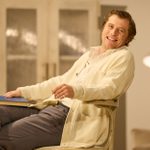 A man (Johnny Flynn) sits in a cane chair, smiling. He wears a pale cardigan and open neck shirt.