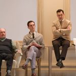 Three men sit in a line against a wall. One sits in an armchair, one slightly higher, sits on a cane chair and the third is highest, sitting on a table. They are all casually dressed in 60s style clothes.