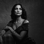 A black and white portrait photo of Tuppence Middleton, wearing heavy bracelets and necklace, sitting on the edge of an arcmchair, looking slightly up and away from the camera.