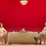 A woman (Tuppence Middleton) and a man (Mark Gattis) sit smiling, opposite each other, at either end of a long table. Both are wearing off-white clothes, the tablecloth is off-white, the floor is pale and the panelled wall behind them is red.