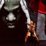 A youg man stands and appears to be practicing fighting. A large image of a man's head, wearing a boxer's hooded gown, fills the wall behind him.