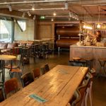 The Understudy bar interior, with a long wooden table with wooden chairs, several round tables - low ones with chairs and tall ones with high stools. Sofas by windows, and a bar with beer taps, and bottles on shelves.