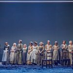 Twelve women stand on a stage in a long line. They are wearing period costumes and all look uncomfortable with the situation they are in, Behind them is a large blank, blue wall, with a white light frame.