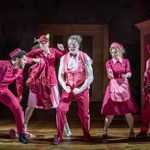 A row of hotel employees, all dressed in red uniforms stand in various poses of anger and rage, and the manin the centre has had his sleeves ripped off.