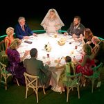 A stage in the round, grass turf of the floor with an illuminated circle. in it is a round table where a group of people are sat around it in a traditional wedding set up.