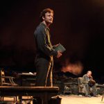 A man stands on a stage, holding a book. In teh background an older man and a woman sit on rustic wooden chairs.