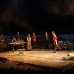 A group of people in simple country clothing stand at one side of an open stage, either side of a man sitting at a long wooden table. They are all looking at two soldiers wearing red jackets and black trousers with a wide red stripe down the side, one of whom is talking.